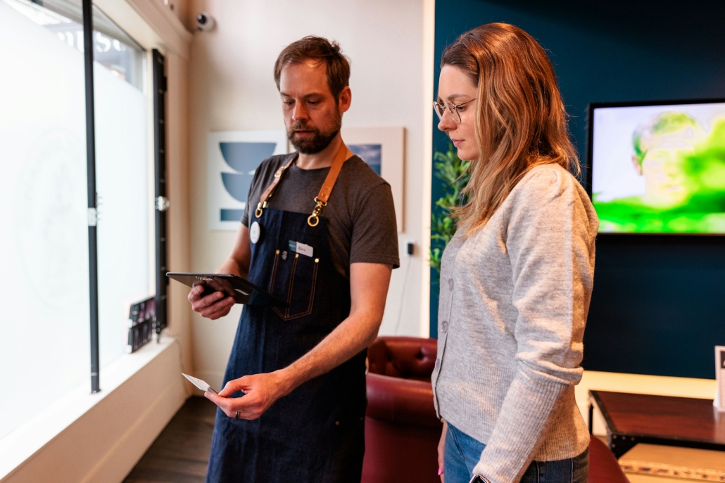 Man holds ipad to scan ID in front of woman who is signing up at a cannabis dispensary. Rules and regulations for safe and legal cannabis consumption help fight historical issues surrounding its access and use. 