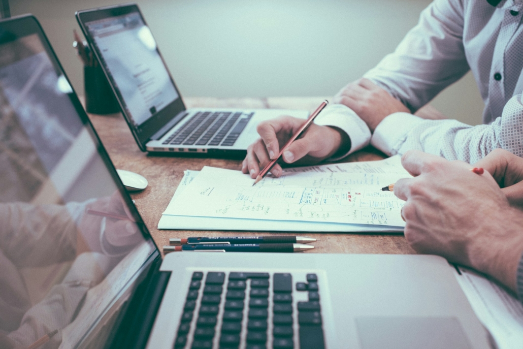 Laptops are on table and the hands of two people are holding pencils and gesturing at paperwork. Cannabis is related to law and regulation.